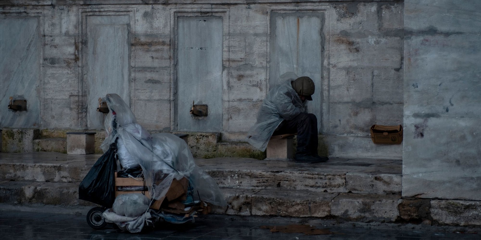 A person sheltering under a plastic sheet on a wet city street, their belongings beside them in a trolley.